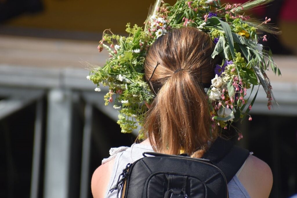 Dziewczyna z plecakiem. Stoi tyłem. Na głowie ma wianek z kwiatów polnych.