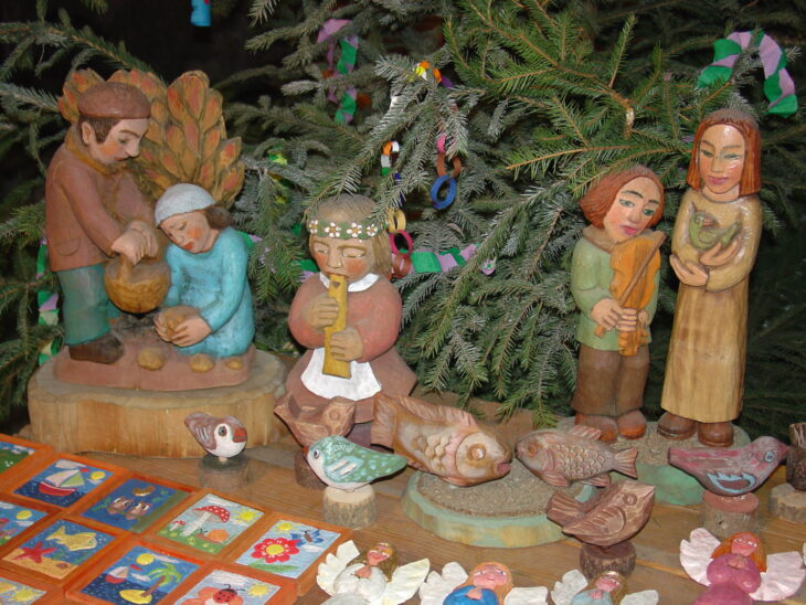 Drewniane rzeźby ustawione na stole. W tle choinka ozdobiona papierowym łańcuchem.