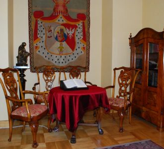 drewniane stare krzesła przy okrągłym stoliku na nim księga a na ścianie sztandar
